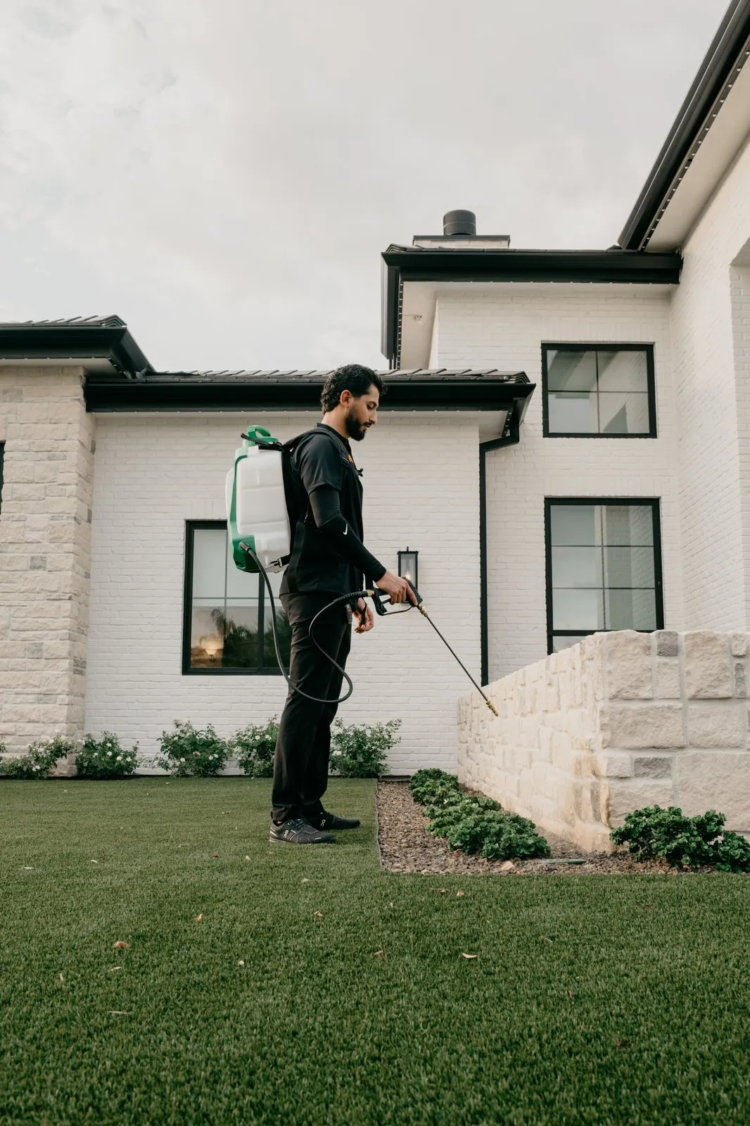Citrus Pest Co technician spraying the base of a stone garden wall along the perimeter of a white brick home
