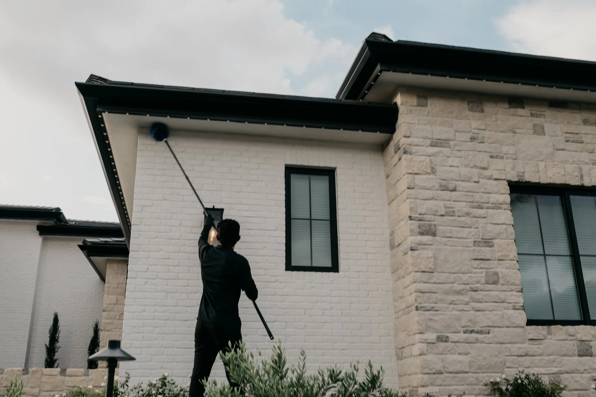 Citrus Pest Co technician using an extension duster to clear webs from the soffit and roofline of a white brick East Valley home