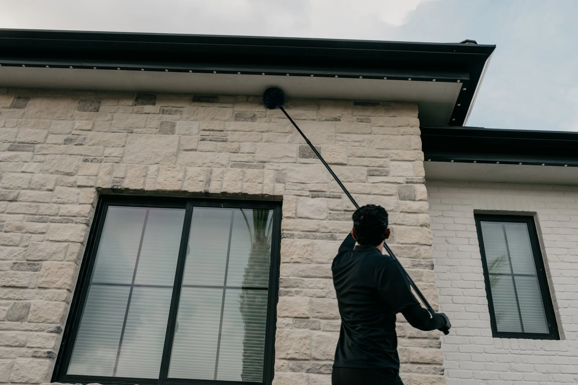 Citrus Pest Co technician using a long extension pole to sweep webs from the soffit and roofline corner of a stone and white brick East Valley home