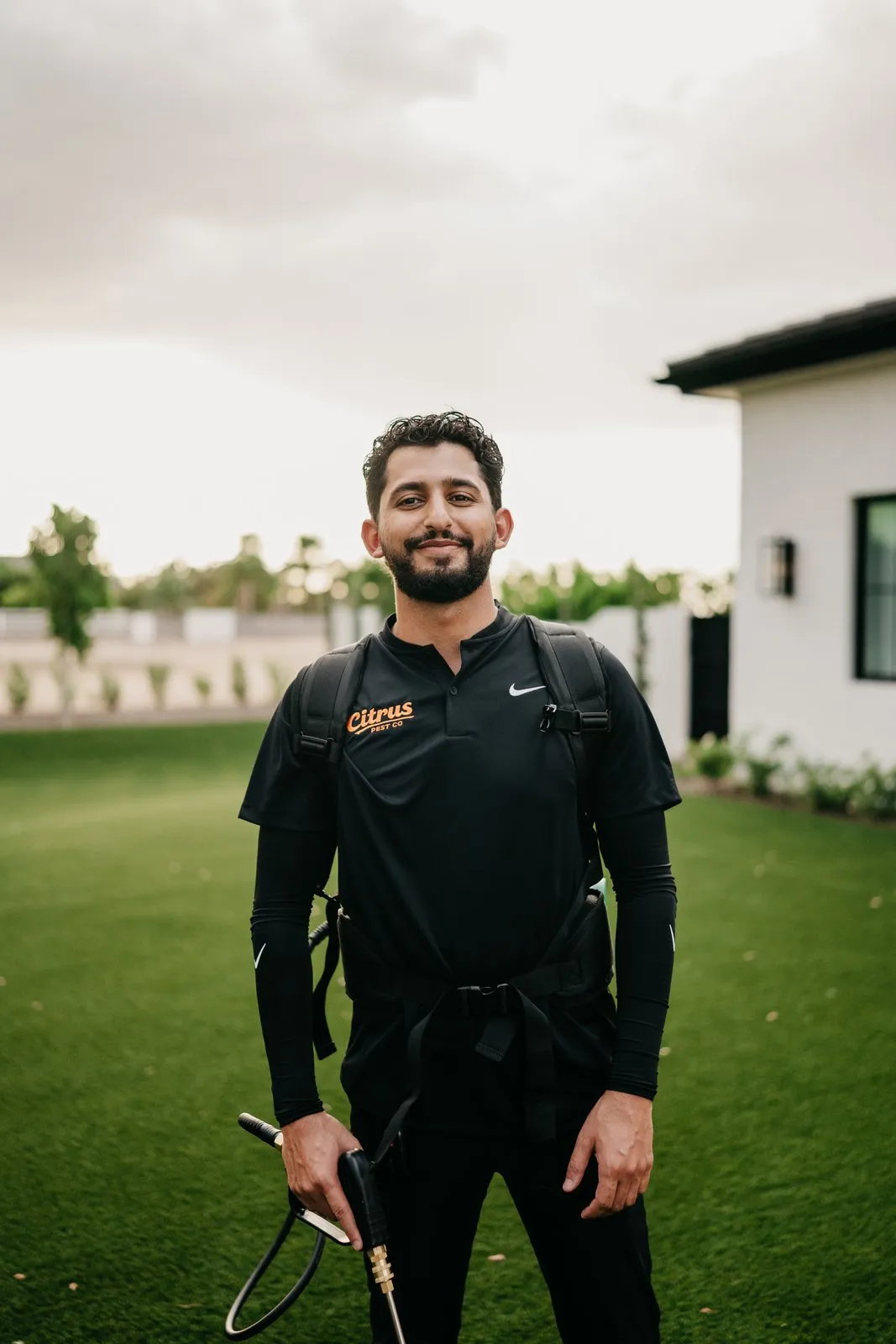 Citrus Pest Co owner in a branded polo, wearing a backpack sprayer on the lawn of an East Valley home