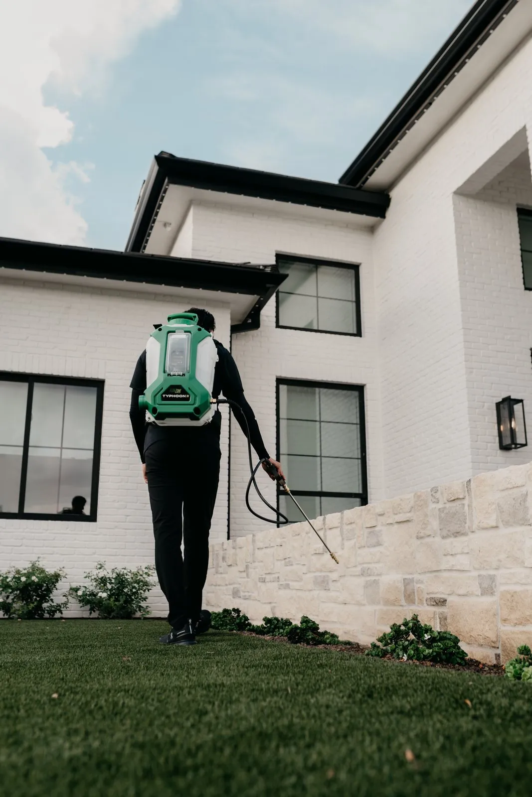Citrus Pest Co technician walking the lawn edge of an East Valley home with a backpack sprayer, treating along the stone wall and foundation
